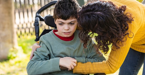 Mamma kramar sin son med funktionsnedsättning som sitter i en rullstol i en park, och visar kärlek och stöd.